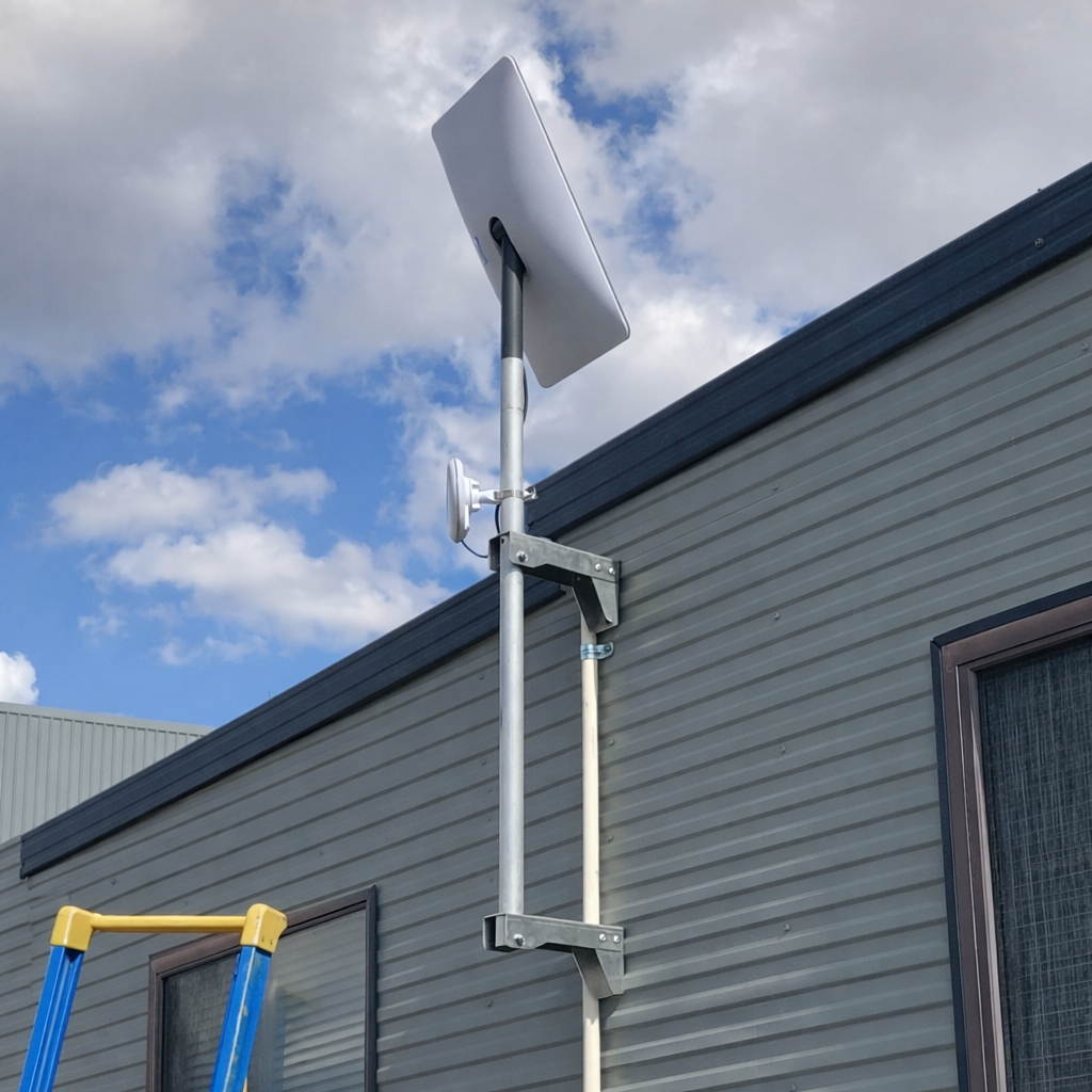 Starlink wall mount pole bracket installed on corrugated building alongside TV antenna under blue sky